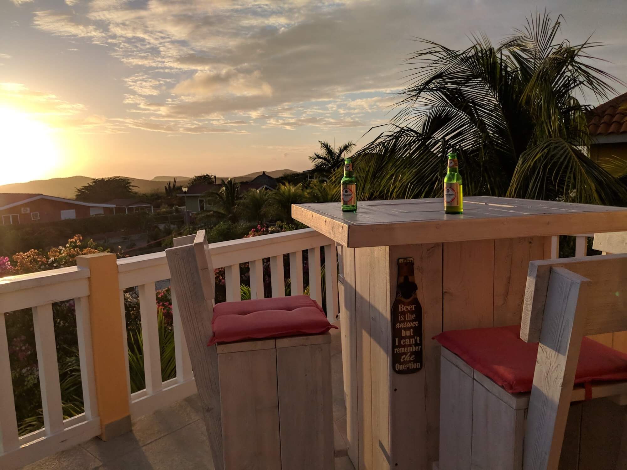 Der hohe Bartisch mit Hockern in der Terrassenecke, zwei Flaschen Bier und Blick auf den Sonnenuntergang